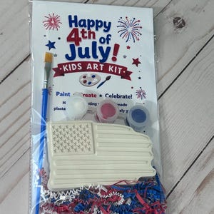 May include: A Happy 4th of July Kids Art Kit in a clear plastic bag containing a white plaster American flag shape, a blue-handled paintbrush, and three small paint pots in white, red, and blue, resting on a bed of red, white, and blue paper shreds.