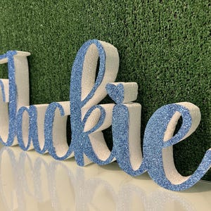May include: A decorative sign spelling out the name "Jackie" in a cursive font. The letters are white with a light blue glitter finish on the front. A small heart shape is above the letter "i". The sign is displayed against a green textured background.