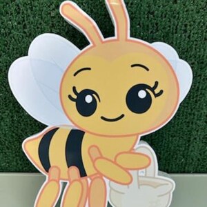 May include: A cartoon-style bee cutout with a yellow body, black stripes, and white wings. The bee is holding a white basket. The image has a green background and a light gray surface.
