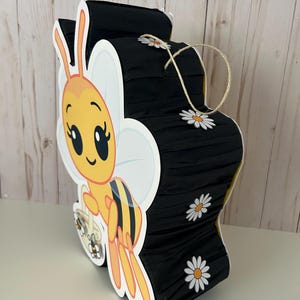 May include: A bee-shaped pi&ntilde;ata with a cartoon bee design. The pi&ntilde;ata is yellow, black, and white, with daisy accents. It has a string for hanging and is made of paper and cardboard.