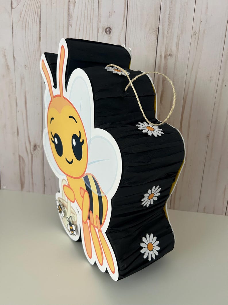 May include: A bee-shaped pi&ntilde;ata with a cartoon bee design. The pi&ntilde;ata is yellow, black, and white, with daisy accents. It has a string for hanging and is made of paper and cardboard.