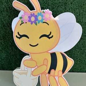 May include: A cheerful, cartoon-style bee cutout with a yellow body, black stripes, and white wings. The bee wears a floral crown and carries a white basket. The letter "D" is visible in the lower right corner.