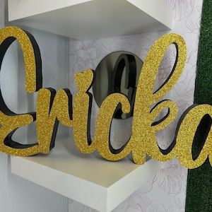 May include: A gold glitter "Erica" name sign with black edging, displayed on a white shelf. The cursive letters are in a decorative font, with a small heart detail above the "i". The sign is a decorative item.