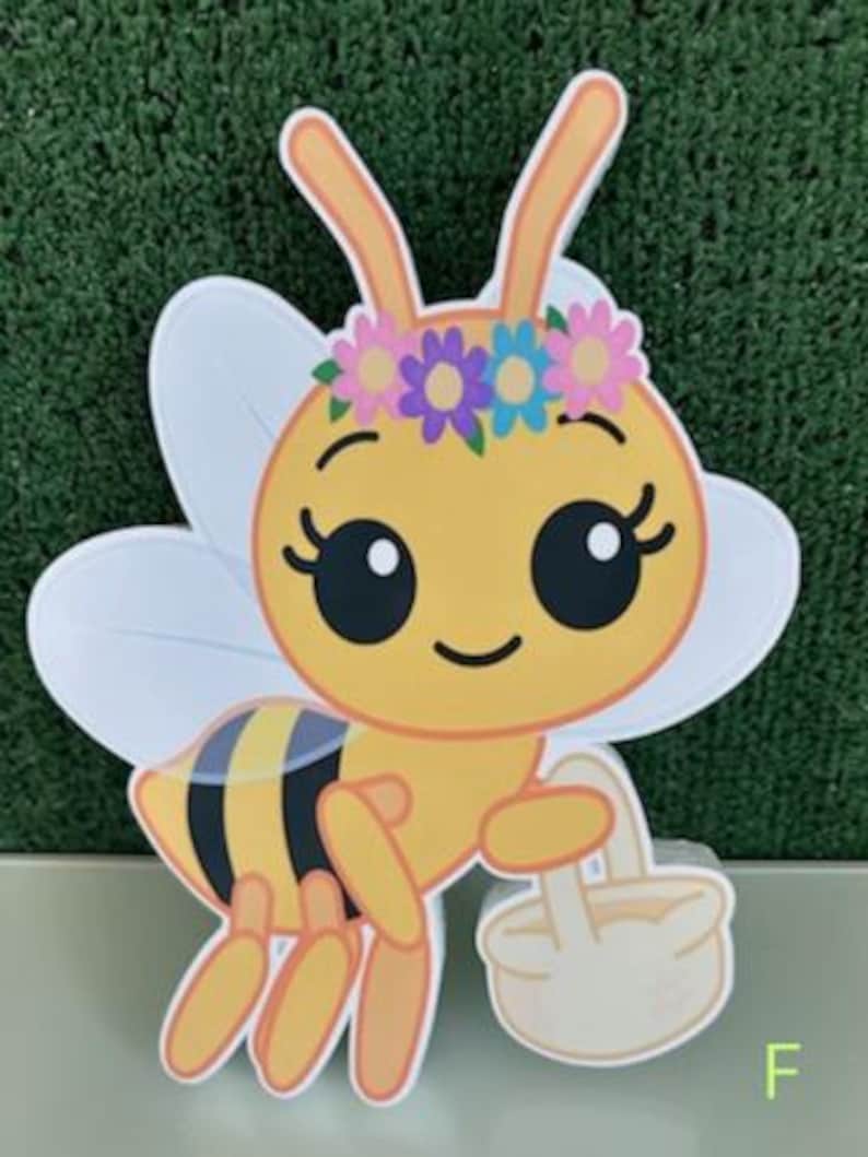 May include: A cartoon bee with a yellow body, black and yellow stripes, and white wings. The bee wears a flower crown and carries a small basket. The letter "F" is in the bottom right corner.