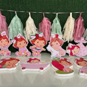 May include: A collection of Strawberry Shortcake-themed cutouts, featuring the character in various poses. The cutouts are in different sizes and shapes, with a pink and white color scheme. A garland of pastel-colored tassels hangs in the background.