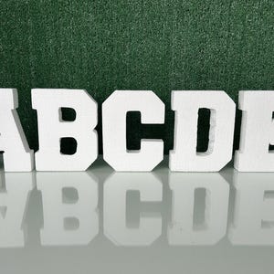 May include: Five white foam letters, A, B, C, D, and E, are arranged in a row. The letters are thick and blocky, with a textured surface. The letters are reflected on a shiny surface, with a green background.