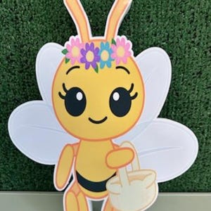 May include: A cheerful, cartoon-style bee cutout with a yellow body, black stripes, and white wings. The bee wears a floral crown and carries a small basket. The letter "E" is visible in the bottom right corner.