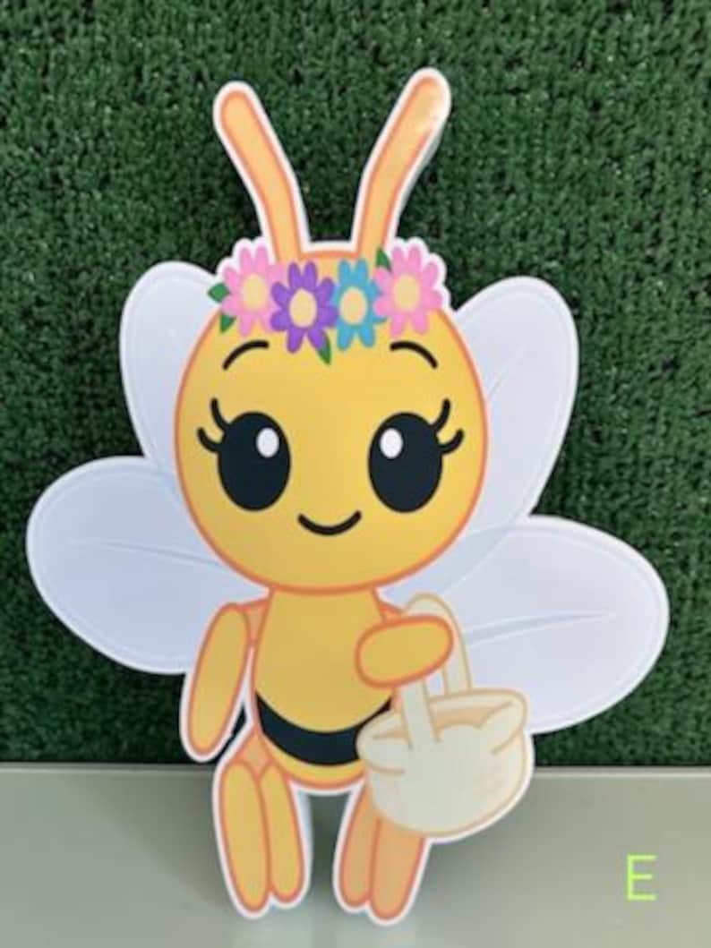 May include: A cheerful, cartoon-style bee cutout with a yellow body, black stripes, and white wings. The bee wears a floral crown and carries a small basket. The letter "E" is visible in the bottom right corner.