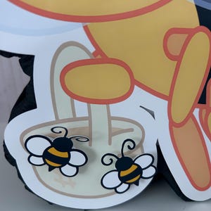 May include: A close-up of a yellow and black bee-themed decoration. Two cartoon bees with white wings and black and yellow striped bodies are featured. The bees are on a white and tan background, with a yellow and orange outline.