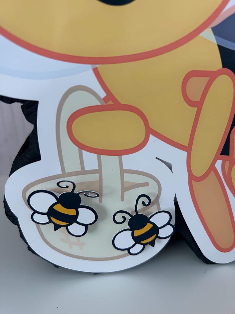 May include: A close-up of a yellow and black bee-themed decoration. Two cartoon bees with white wings and black and yellow striped bodies are featured. The bees are on a white and tan background, with a yellow and orange outline.