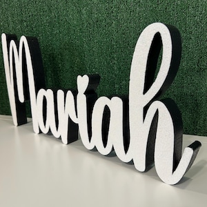 May include: A decorative sign spelling "Mariah" in a cursive font. The letters are white with glitter, outlined in black. A small heart is above the "i". The sign is on a white surface with a green background.