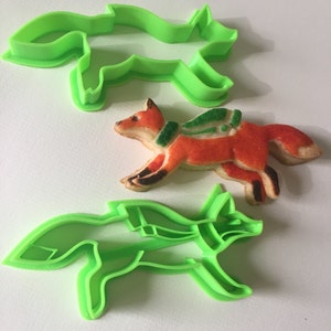 Puede incluir: Dos cortadores de galletas de zorro de plástico verde y una galleta de zorro decorada. La galleta es naranja con glaseado verde y blanco.