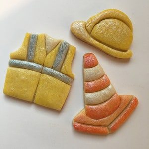 May include: Three yellow and orange sugar cookies decorated with icing. The cookies are shaped like a hard hat, a safety vest, and a traffic cone.