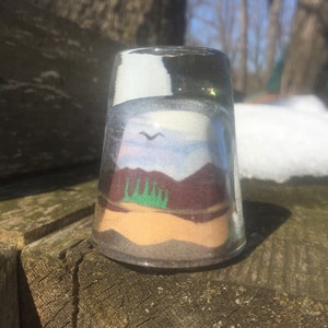 May include: A glass jar with a layered sand art scene depicting a mountain range, a sky with a bird flying, and a beach with green trees.