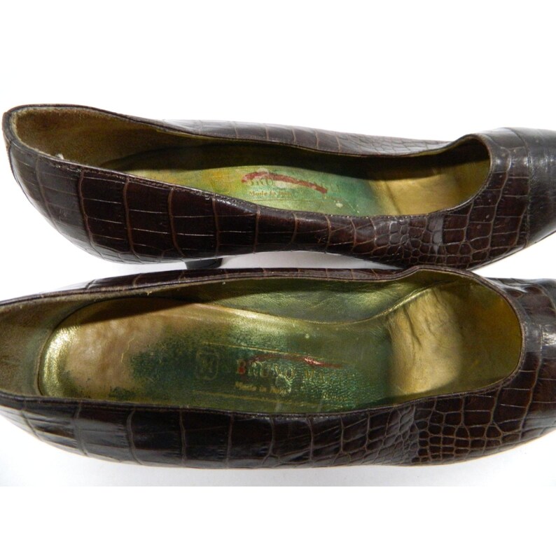 Op de afbeelding: Een paar bruine, getextureerde leren ballerina's. De schoenen hebben een spitse neus en een lage hak. De binnenkant van de schoen is goudkleurig met een groen en rood label. De schoenen worden op een witte achtergrond gepresenteerd.
