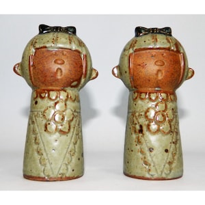 May include: A pair of ceramic salt and pepper shakers shaped like girls with brown faces, green dresses, and black bows in their hair.