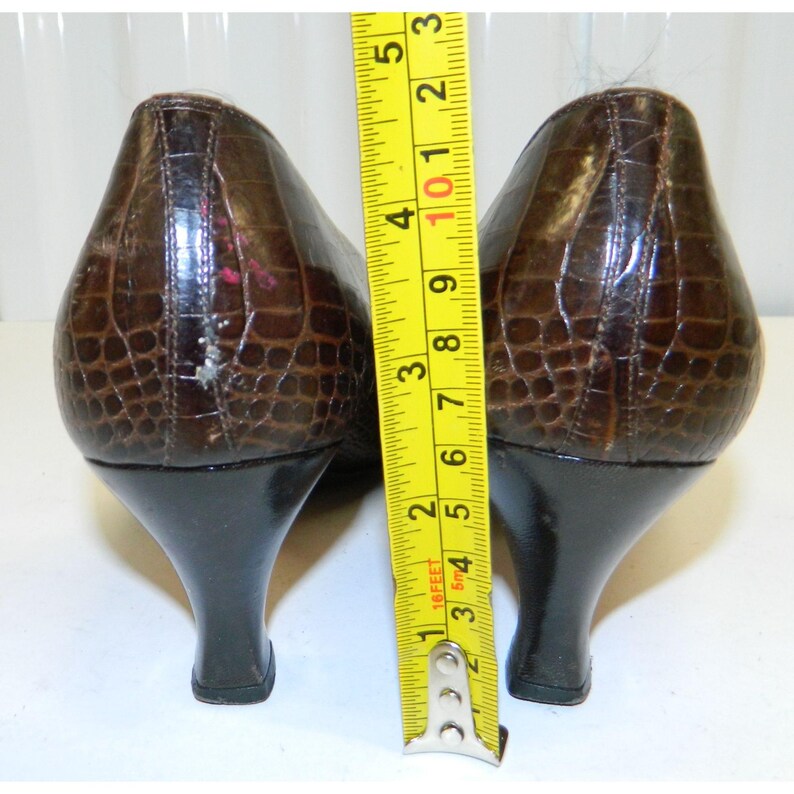 Op de afbeelding: Een paar bruine, getextureerde leren pumps met zwarte hakken. De hakken zijn ongeveer 7,6 cm hoog. De schoenen hebben een spitse neus en een reptielachtig patroon. Een gele meetlint is naast de schoenen geplaatst.