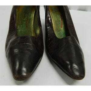 Op de afbeelding: Een paar bruine leren pumps met een textuur die op krokodillenleer lijkt. De schoenen hebben een spitse neus en een lage hak. De binnenkant van de schoen is groen en goud.