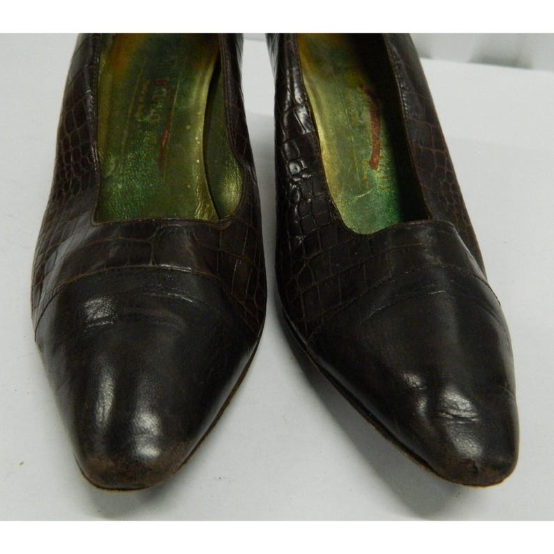 Op de afbeelding: Een paar bruine leren pumps met een textuur die op krokodillenleer lijkt. De schoenen hebben een spitse neus en een lage hak. De binnenkant van de schoen is groen en goud.