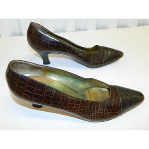 Op de afbeelding: Een paar bruine leren pumps met een imitatie krokodillenleer textuur. De schoenen hebben een spitse neus en een kleine, zwarte hak. De binnenkant van de schoen is lichtgroen.