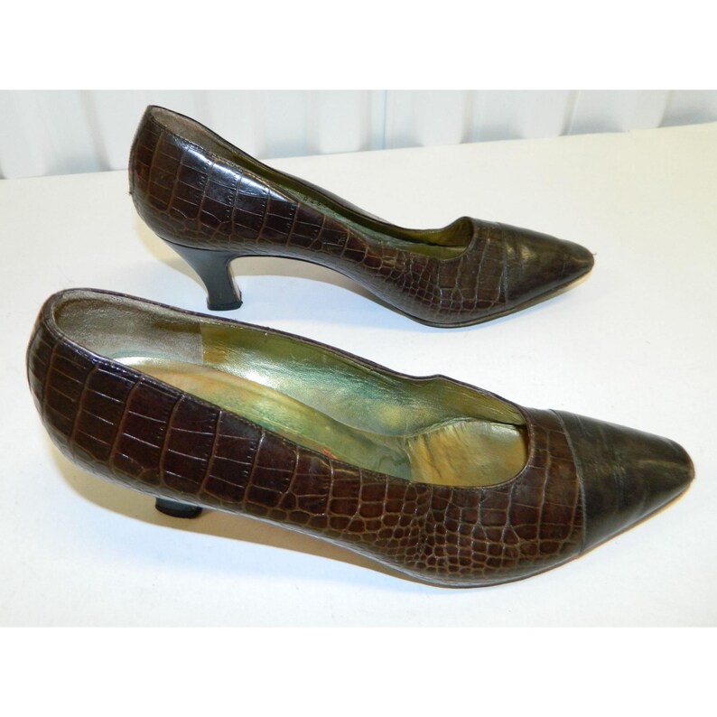 Op de afbeelding: Een paar bruine leren pumps met een imitatie krokodillenleer textuur. De schoenen hebben een spitse neus en een kleine, zwarte hak. De binnenkant van de schoen is lichtgroen.
