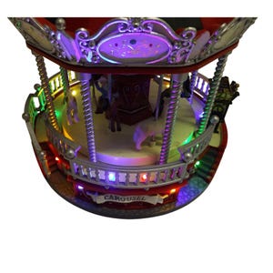 May include: A miniature carousel with a red and silver frame, featuring white horses and figures on a rotating platform. The structure is illuminated by multi-colored LED lights along the railings and roof, with the word CAROUSEL printed on a banner.