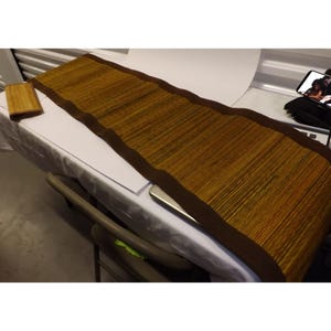 May include: A long, rectangular brown woven table runner with a dark brown border. A small, matching square coaster is to the left. The runner is made of natural fibers and has a textured appearance.