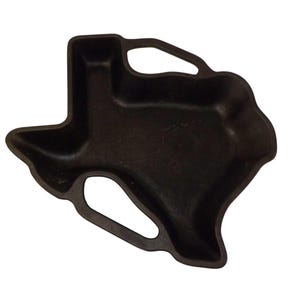 May include: A black cast iron serving dish in the shape of the state of Texas. The dish has a textured surface and two handles. This decorative piece is suitable for serving appetizers or small dishes.