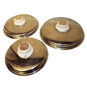May include: Three vintage, round, metal hot water bottle caps. Each cap has a white plastic screw-on top with a gold-colored center. The caps are made of shiny, silver-colored metal and are arranged on a white background.