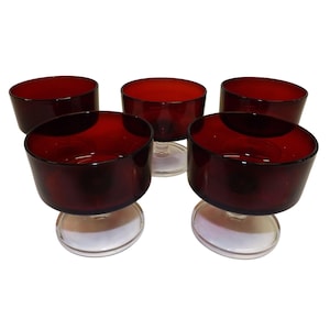 May include: Five ruby red glass dessert bowls. Each bowl has a rounded, angled shape and sits atop a clear, circular base. The bowls are arranged in a slightly offset pattern against a white background.