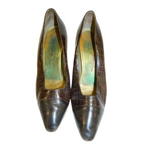Op de afbeelding: Een paar bruine leren pumps met een krokodillenstructuur. De neuzen zijn donkerder bruin. De binnenkant van de schoenen heeft een goud- en groentint.