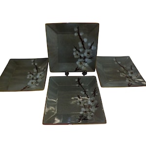 May include: Set of four square ceramic plates. Each plate is a muted gray-green with a floral design of white blossoms and dark brown branches. The plates are arranged on a white background.