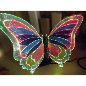 May include: A decorative butterfly lamp featuring translucent, multi-colored wings with glowing fiber optic edges. The wings display segments of red, blue, and green, while the central body is dark and textured. It sits on a black base.