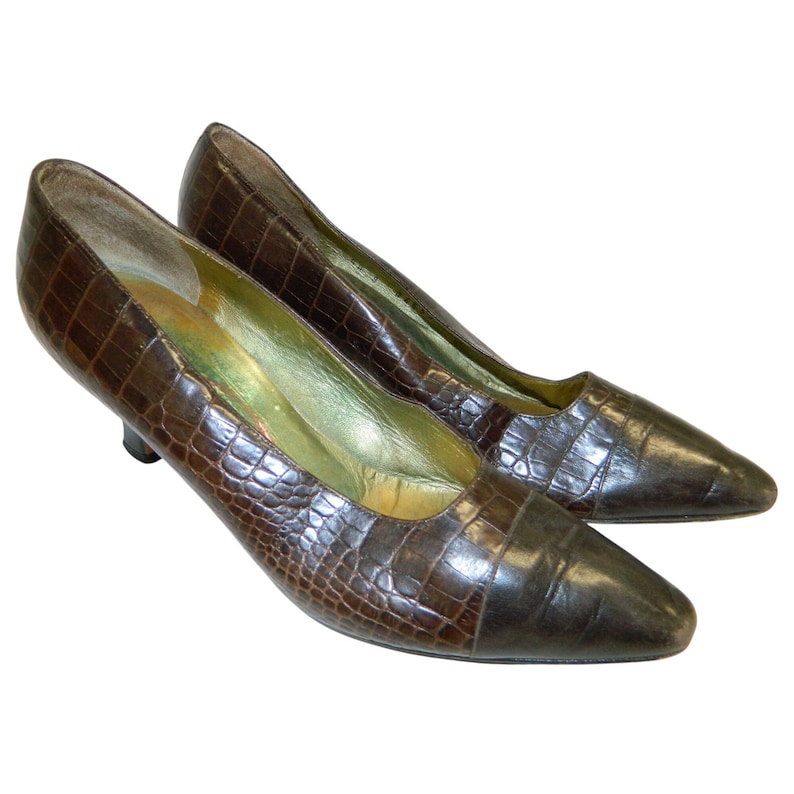Op de afbeelding: Een paar bruine pumps met lage hakken met een textuur van imitatiekrokodillenleer. De schoenen hebben spitse tenen en een donkerbruine kleur op de neus. De binnenkant van de schoen is lichtgoud van kleur.
