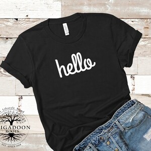 May include: Black t-shirt with the word "hello" in white script. The shirt is folded, with the sleeves cuffed. The bottom of the shirt is paired with blue denim jeans. The shirt has a tag that says "Canvas".