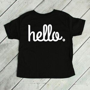 May include: Black short-sleeved t-shirt with the word "hello" in white script across the front. The word is slightly curved and has a small dot at the end. The shirt is laying on a light wood surface.