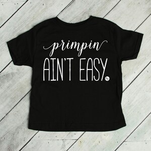 May include: Black t-shirt with the white text "primpin AIN'T EASY." The text is in a mix of cursive and block letters. The shirt is displayed on a light wood grain surface.