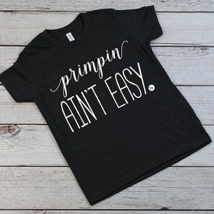 May include: Dark gray t-shirt with the phrase "primpin ain't easy" in white script and block letters. The shirt is displayed on a light wood-grain surface.