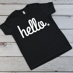以下が含まれることがあります： 白の筆記体で「hello」と書かれた濃い灰色のTシャツ。柔らかい素材でできており、カジュアルウェアに適しています。「hello」の文字が主なデザイン要素です。