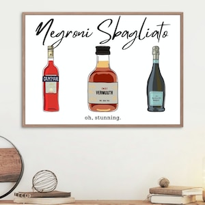 Op de afbeelding: Een witte ingelijste print met de tekst "Negroni Sbagliato" in zwarte cursieve letters. De print toont drie flessen alcohol: een rode fles met het label "Campari", een bruine fles met het label "Sweet Vermouth" en een blauwe fles met het label "Prosecco". De tekst "oh, stunning." is onder de flessen gedrukt.