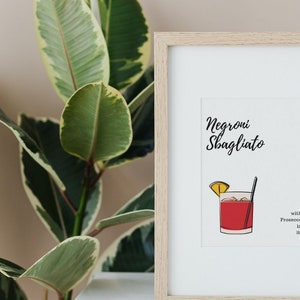 Op de afbeelding: Een ingelijst print met een witte achtergrond en zwarte tekst die "Negroni Sbagliato with Prosecco in it" luidt. De print toont een cartoonillustratie van een rode cocktail in een glas met een citroenwig en een rietje.