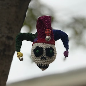 May include: A crocheted skull wearing a colorful jester hat with bells. The skull has green eyes and a white face.