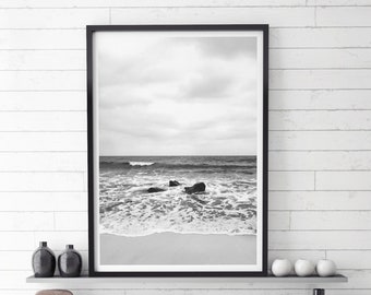 May include: Black and white photograph of a beach scene with waves crashing on the shore. The photo is framed in a black frame and is hanging on a white wall.