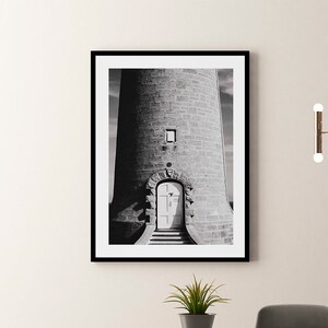 Puede incluir: Impresión en blanco y negro enmarcada de un faro de piedra con una puerta blanca. La impresión se muestra encima de una mesa de comedor de madera oscura con sillas grises y una pequeña planta en maceta. Un candelabro moderno cuelga en el fondo.