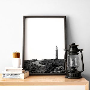 Puede incluir: Fotografía en blanco y negro de un faro en una costa rocosa. La foto está enmarcada en un marco negro y se exhibe en un estante de madera con una linterna negra y una pila de libros.