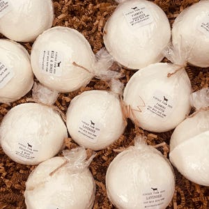 Op de afbeelding: Een verzameling witte badbruisballen, elk verpakt in doorzichtig cellofaan en vastgebonden met touw. De badbruisballen hebben labels met tekst zoals "Vintage Farm Suds" en verschillende geuren zoals "Lavendel" en "Melk & Honing". Ze liggen op een bed van bruin papier.
