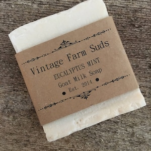 May include: A bar of white goat milk soap wrapped in brown paper with the text "Vintage Farm Suds Eucalyptus Mint Goat Milk Soap Est. 2014" printed on the label.