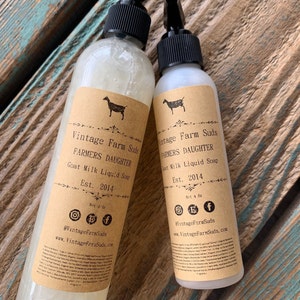 Könnte beinhalten: Zwei transparente Flaschen Ziegenmilch-Flüssigseife mit schwarzen Sprühköpfen. Die Etiketten sind beige mit einer Ziegensilhouette und dem Text "Vintage Farm Suds, FARMERS DAUGHTER, Est. 2014". Auf einer verwitterten Holzoberfläche.