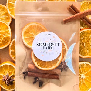 May include: A clear bag filled with dried orange slices, cinnamon sticks, cloves, and a decorative iridescent accent. The bag has a round label that reads "SOMERSET FARM EST. 2025". The background features more dried orange slices.