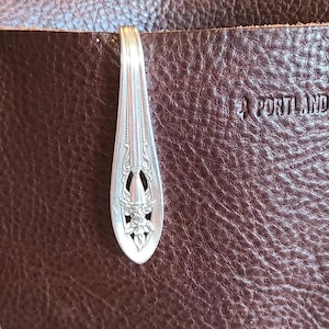 May include: Close-up of a brown leather bag with a silver spoon clip. The bag has a textured surface and a button that says "Leather Goods". The text "Portland Leather" is visible on the bag.